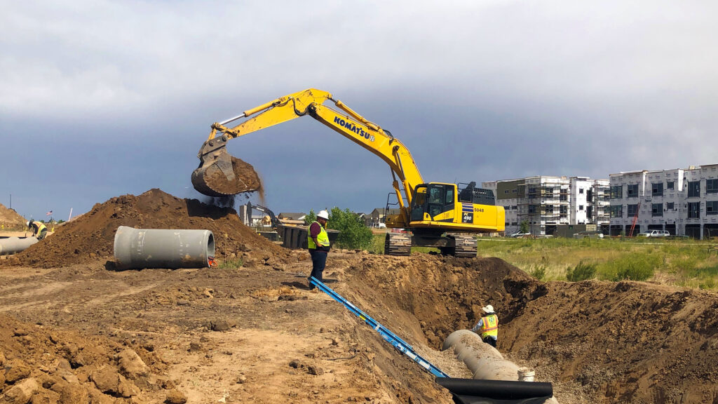 Excavator dumping dirt near a ditch with workers laying pipe